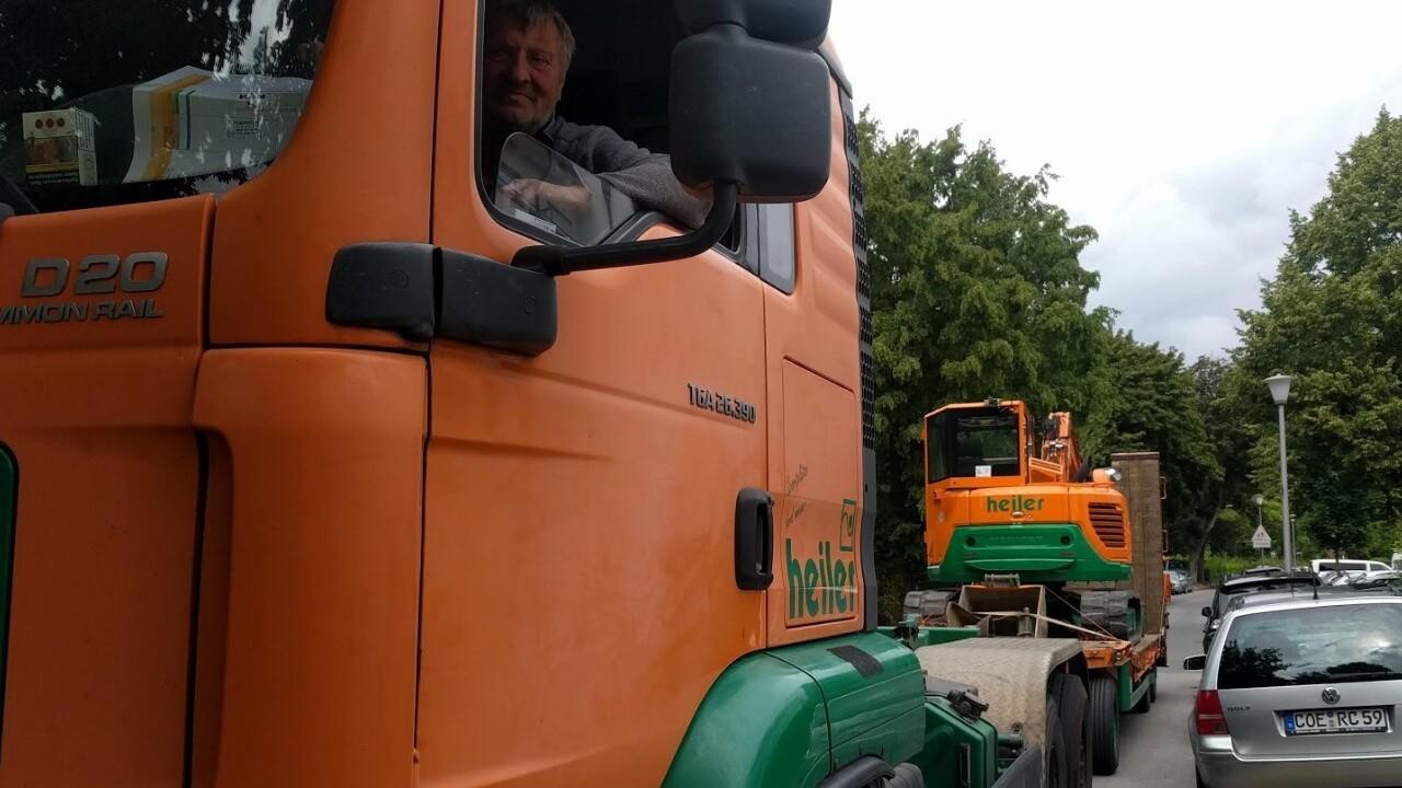 Lkw mit orangefarbener Kabine und Fahrer auf einer Straße, im Hintergrund eine orange Baustellenmaschine.