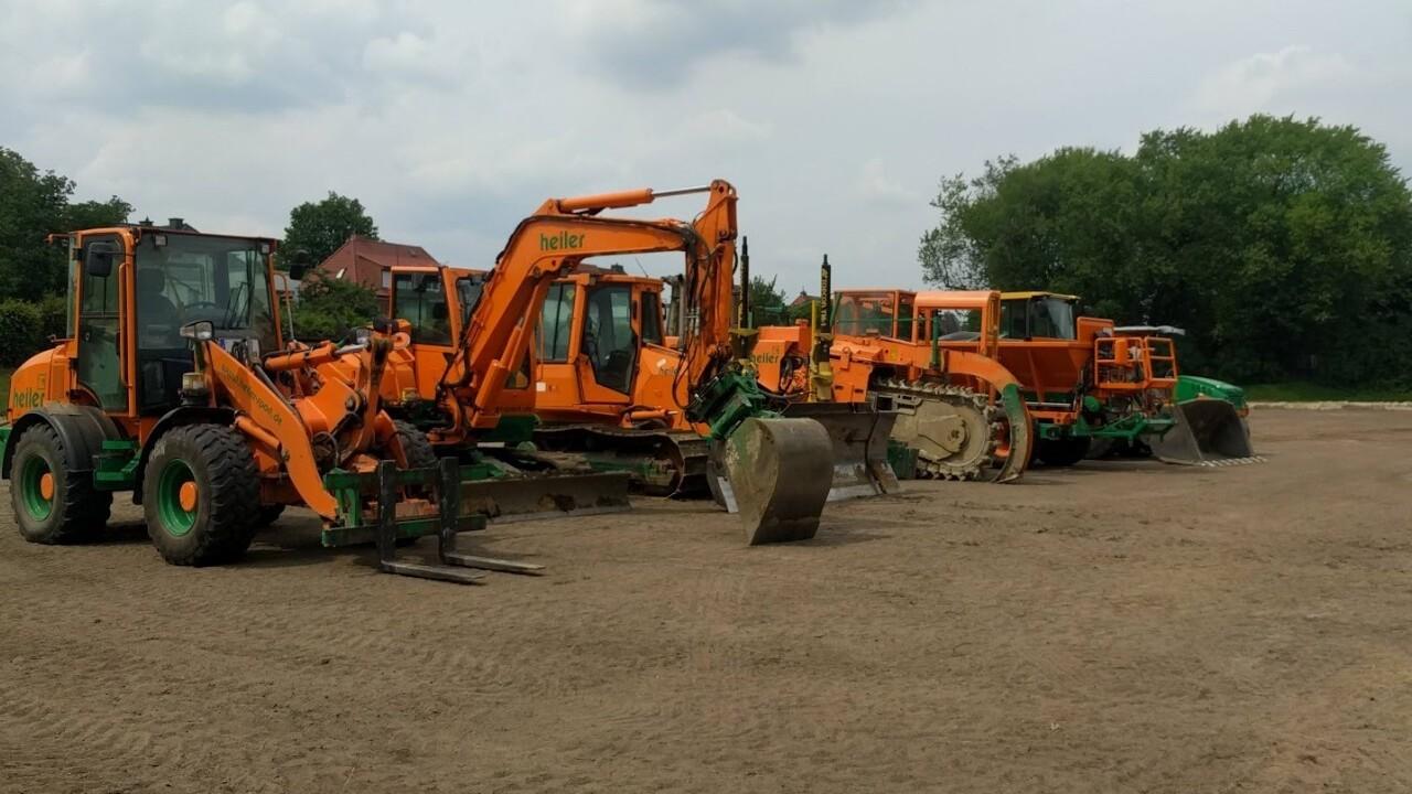 Reihe orangefarbener Baumaschinen auf einer unbefestigten Fläche unter einem wolkigen Himmel.
