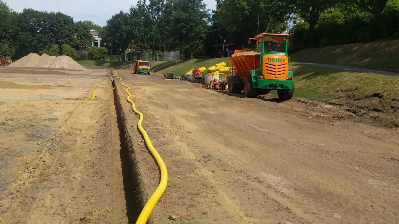 Baustelle mit gelbem Schlauch, Maschinen und Erde; im Hintergrund Bäume und unebene Fläche für Arbeiten.