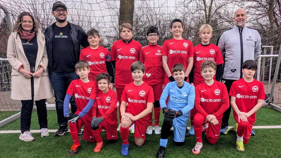 Gruppensfoto einer Jugendfußballmannschaft in roten Trikots mit Betreuern auf einem Sportplatz.