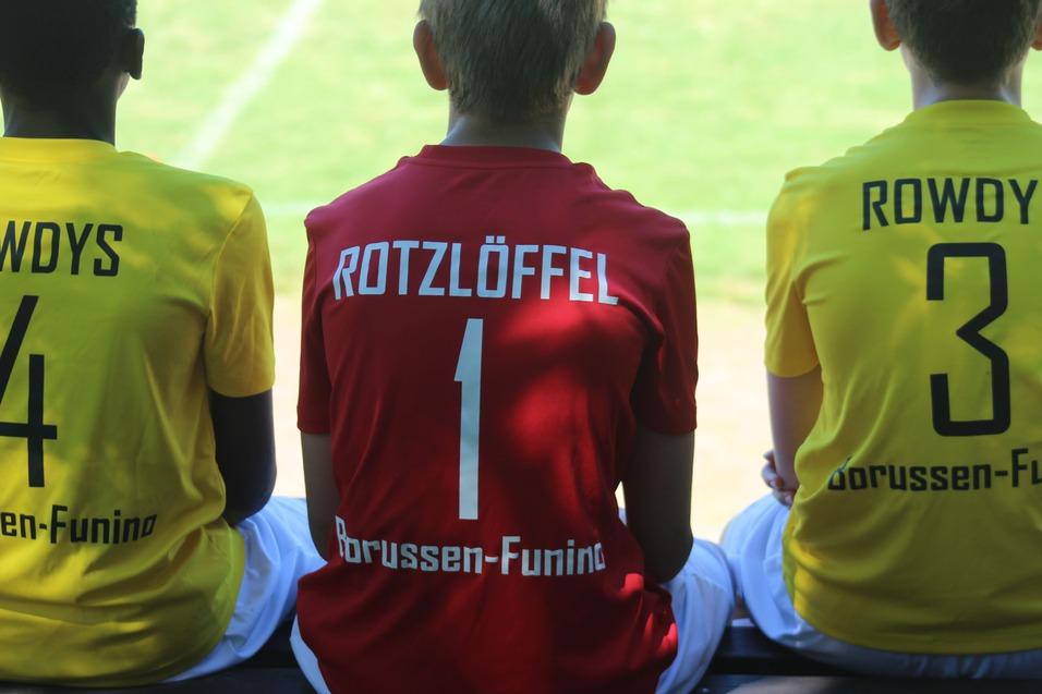 Drei Kinder in Trikots sitzen nebeneinander auf einer Bank, mit den Rückennummern 4, 1 und 3.