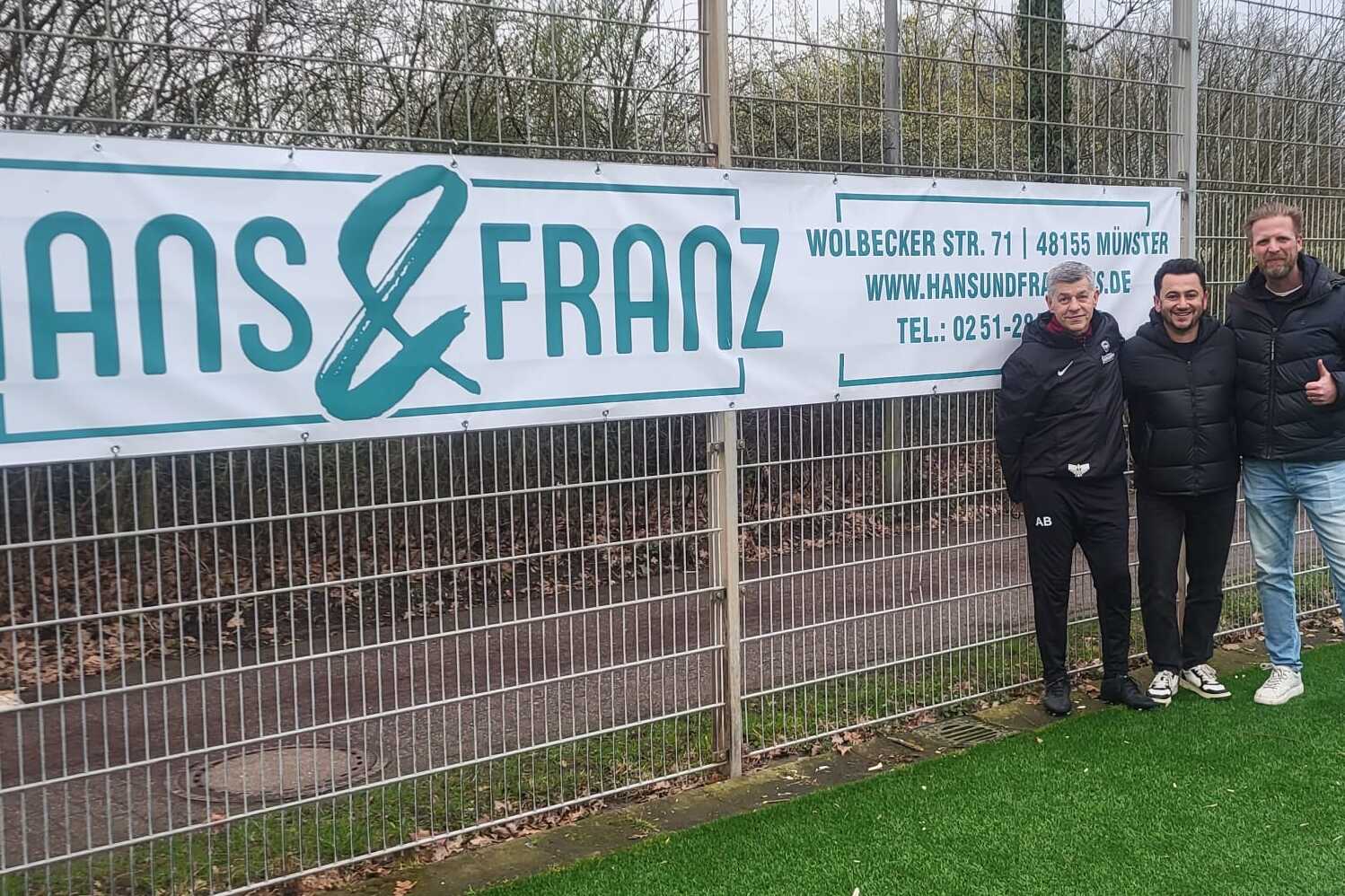 Drei Personen stehen vor einem Zaun mit einem Banner von "HANS & FRANZ" auf einem Fußballplatz.