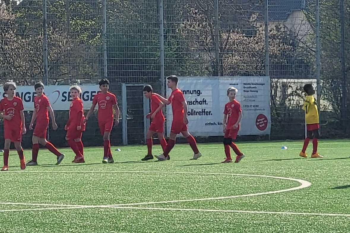 Sechs Fußballspieler in roten Trikots laufen auf einem Kunstrasenplatz, während ein Spieler in blauem Trikot wartet.