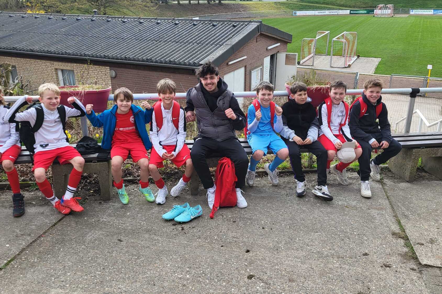 Gruppe von zehn jungen Fußballspielern, sitzend auf einer Bank, im Hintergrund ein Fußballfeld und ein Sportgebäude.