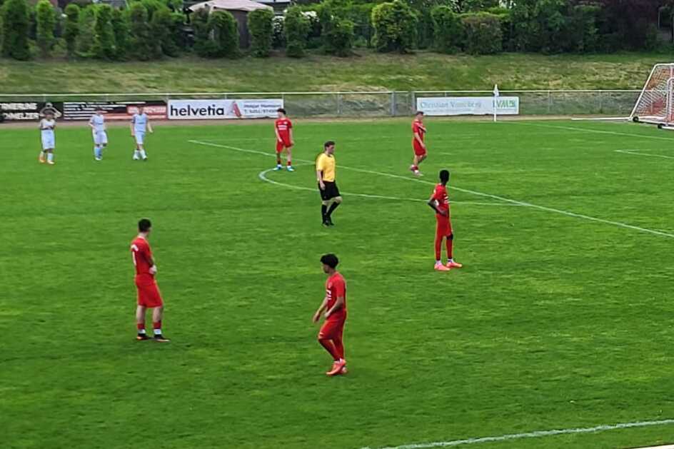 Fußballspiel auf einem Rasenplatz mit Spielern in roten Trikots, Referee und Tore im Hintergrund.