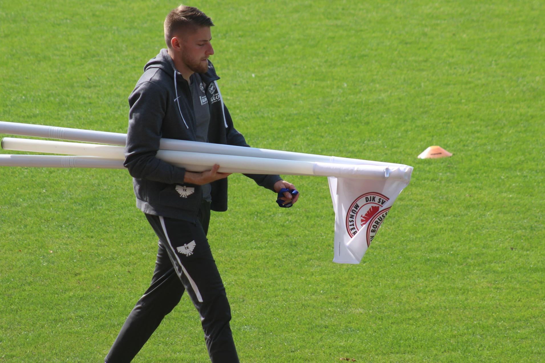 Mann trägt mehrere weiße Rohre und eine Flagge über den grünen Fußballplatz.