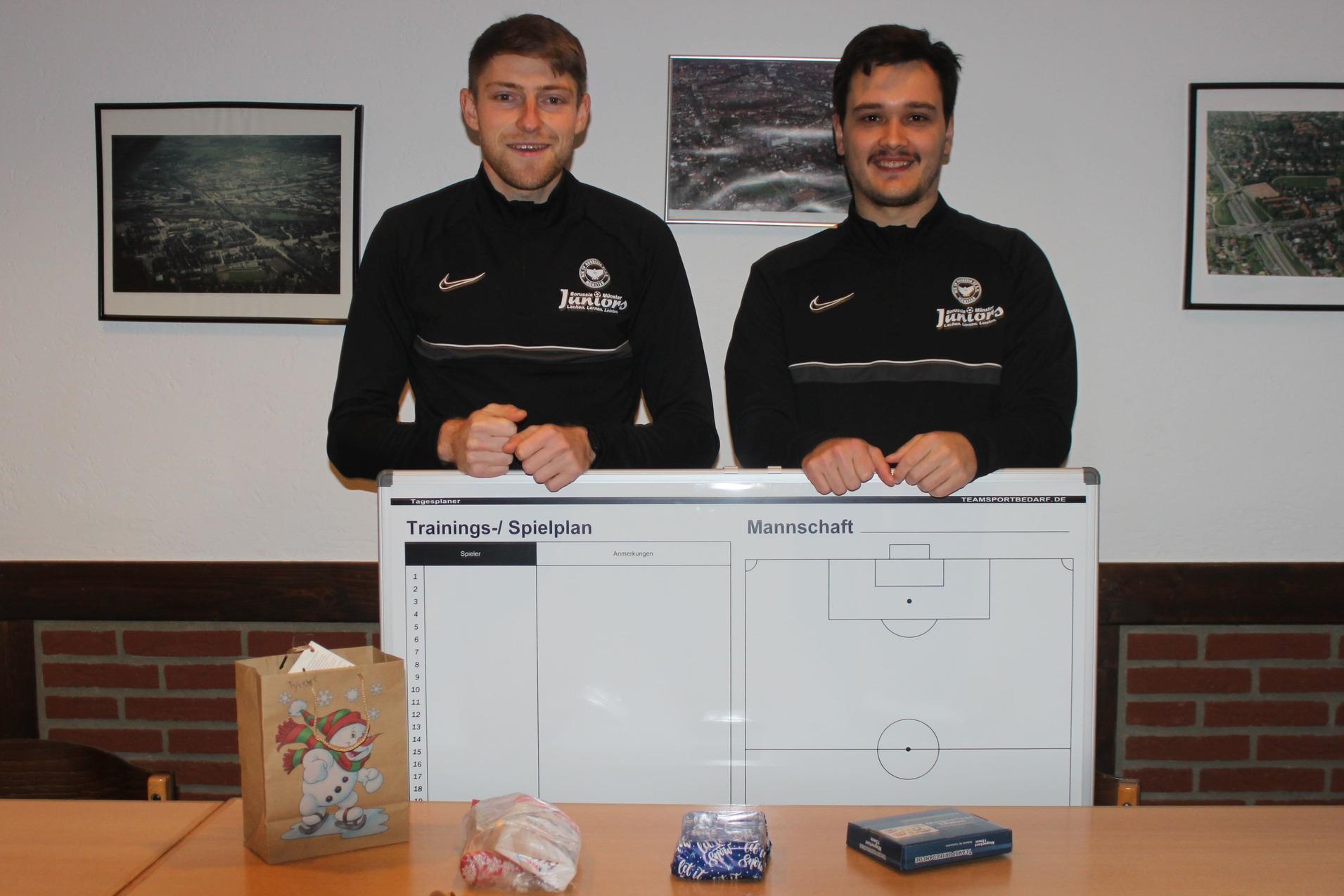 Zwei Fußballtrainer stehen hinter einem großen Trainingsspielplan, während Geschenke und Süßigkeiten auf dem Tisch liegen.