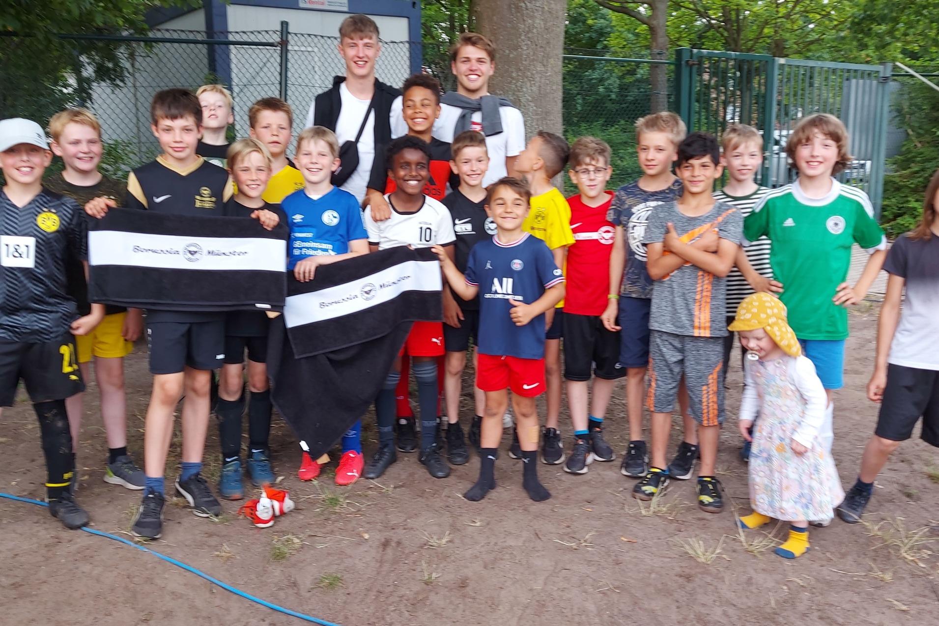 Gruppe von 18 Jungen in Fußballkleidung posiert zusammen im Freien, einige halten Banner.