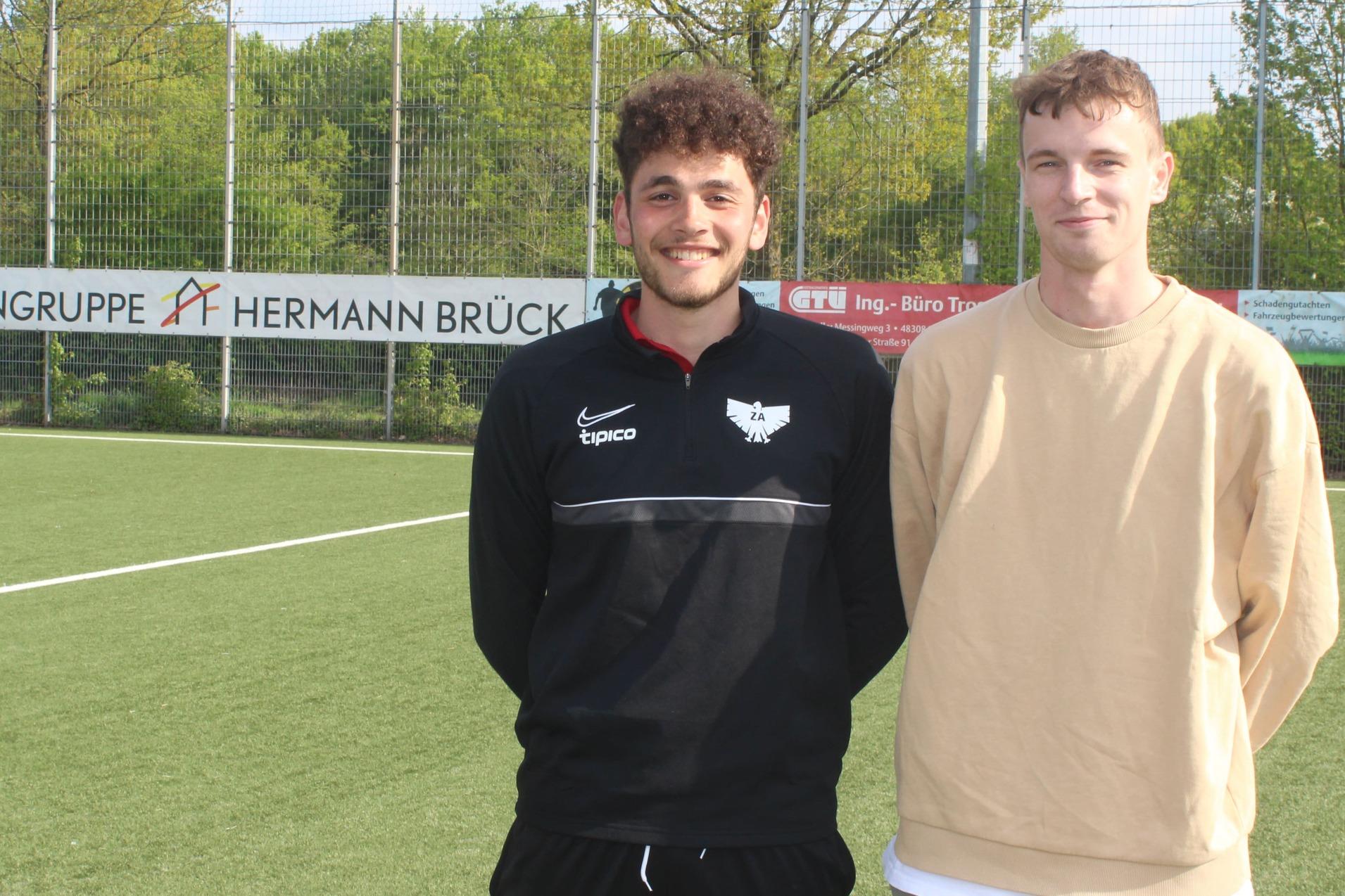 Zwei junge Männer stehen lächelnd auf einem Fußballfeld, im Hintergrund sind Sponsorenbanner und Bäume zu sehen.