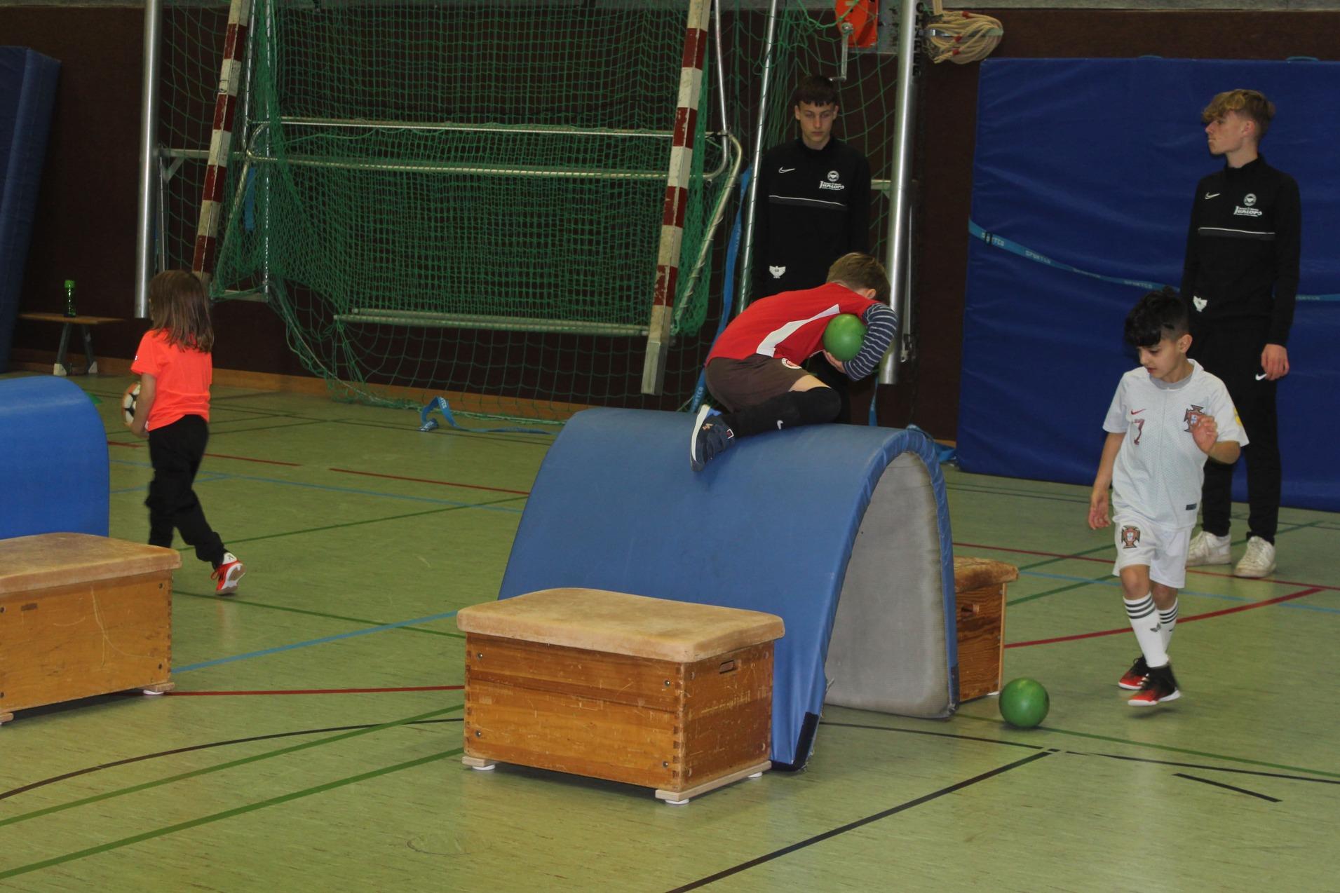 Kinder spielen in einer Sporthalle, während zwei in Trainingsanzügen zuschauen. Eine Person springt über ein Hindernis.