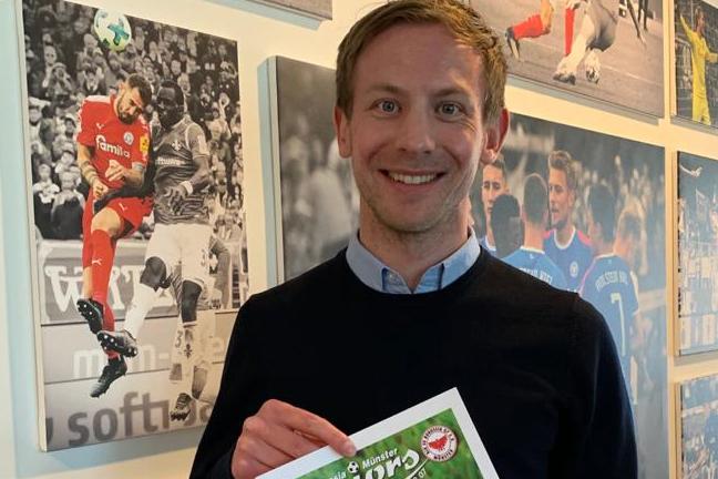 Mann steht vor einer Wand mit Fußballfotos und hält ein Plakat der Borussia & Münster Juniors in der Hand.