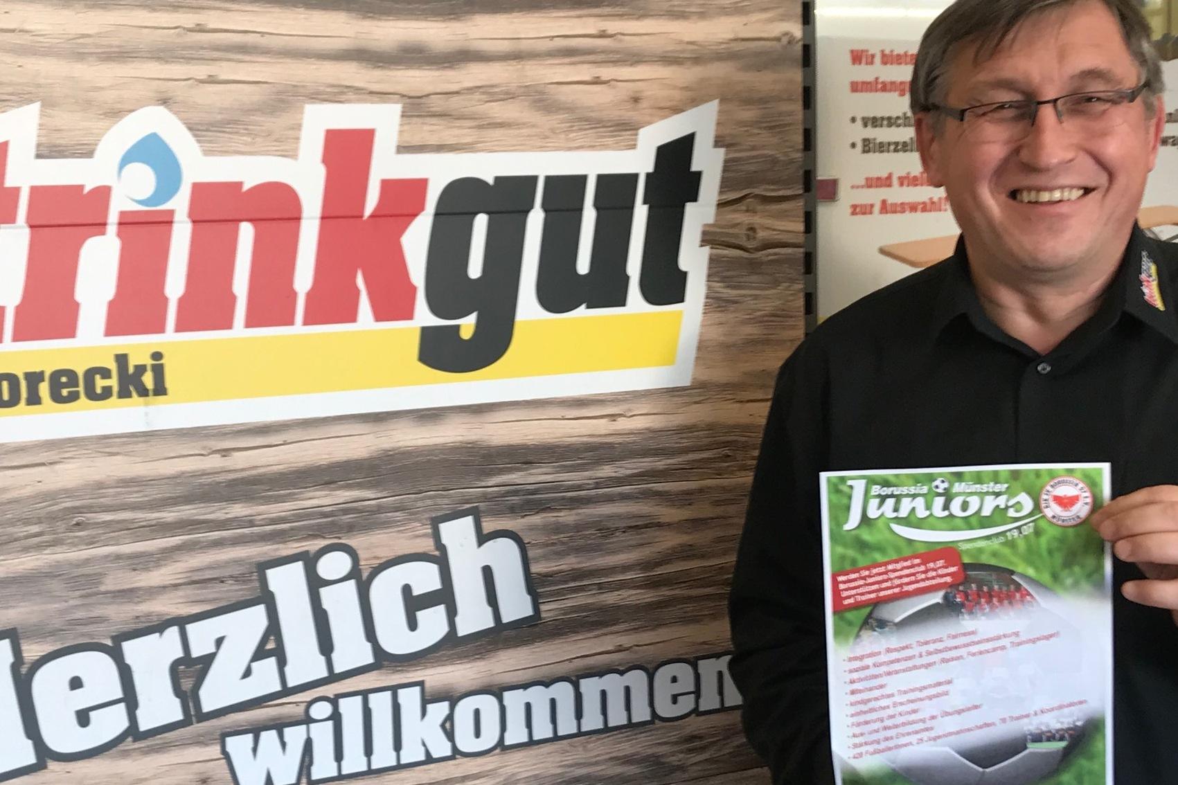 Mann hält ein Informationsblatt vor einem plakatierten Hintergrund mit der Aufschrift „trinkgut Gorecki“ und „Herzlich willkommen“.