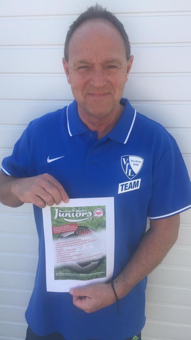 Mann in blauem Team-Trikot hält ein Informationsblatt über eine Juniorenveranstaltung vor einer hellen Wand.