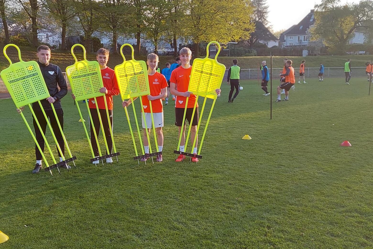 Fünf Kinder in Sportkleidung halten leuchtend gelbe Trainingspuppen auf einem Fußballplatz bei Sonnenuntergang.