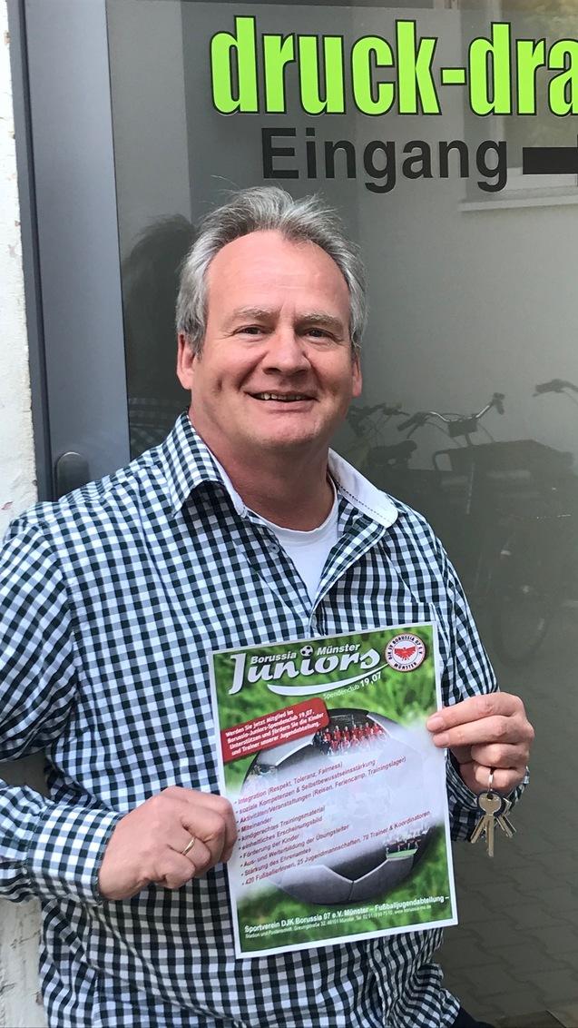 Mann mit grauem Haar hält ein Plakat vor einem Eingang mit der Aufschrift „druck-drauf“. Fahrräder sind im Hintergrund sichtbar.
