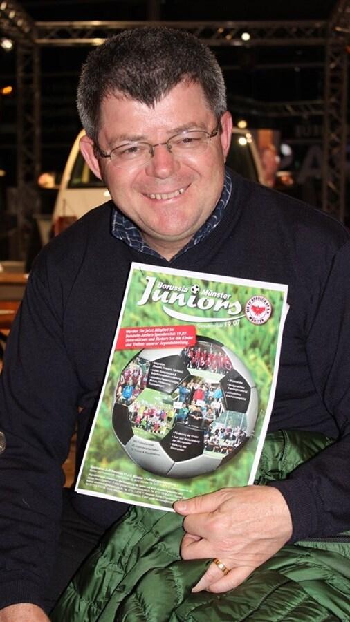 Mann mit Brille hält eine Broschüre von Borussia Münster Juniors in der Hand, lächelnd vor einer Holzplatte.