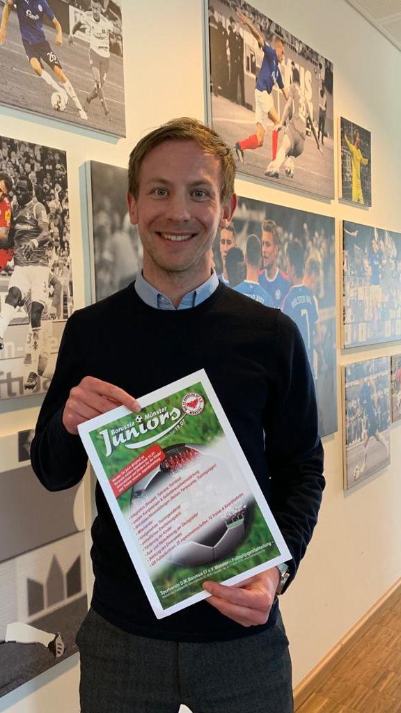 Mann steht vor einer Wand mit Fußballfotos und hält ein Plakat der Borussia & Münster Juniors in der Hand.