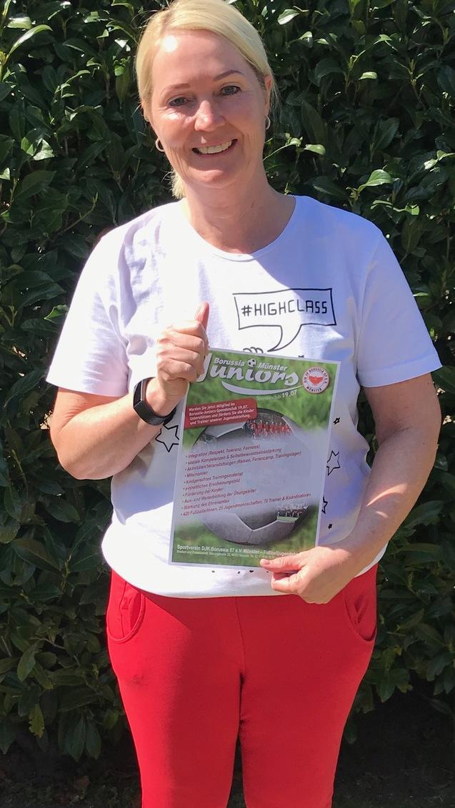 Frau in weißem T-Shirt und roten Hosen hält ein Plakat mit Informationen, vor einer Hecke im Freien.