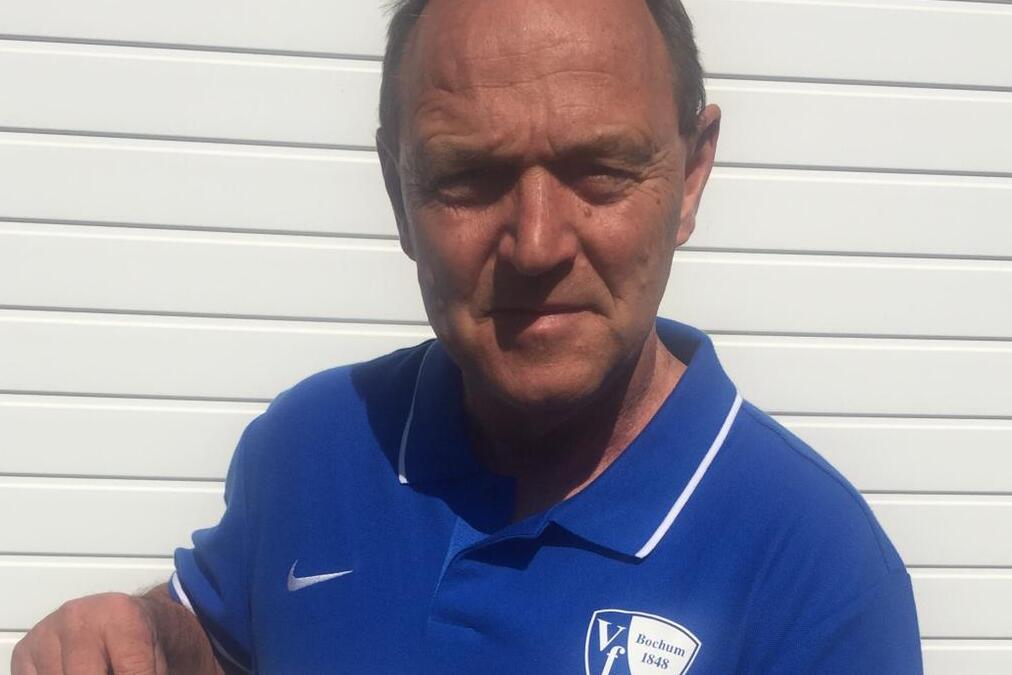Mann in blauer Sportkleidung hält ein Informationsblatt über ein Fußballprogramm für Jugendliche vor einer weißen Wand.