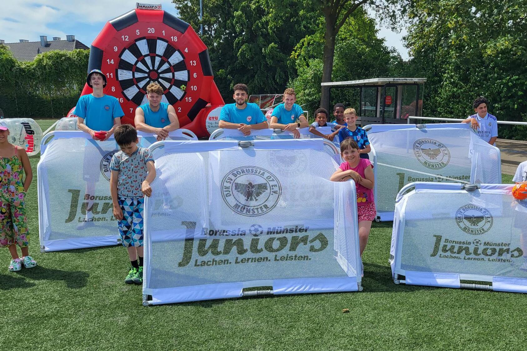 Gruppe von Kindern und Betreuern auf einem Sportplatz hinter transparenten Spielschildern mit der Aufschrift „Juniors“.