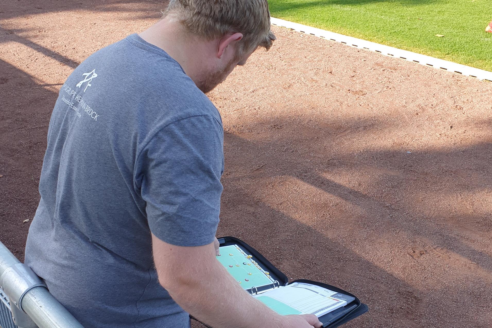 Ein Trainer beobachtet ein Fußballspiel und notiert auf einem Tablet, während Spieler auf dem Feld trainieren.