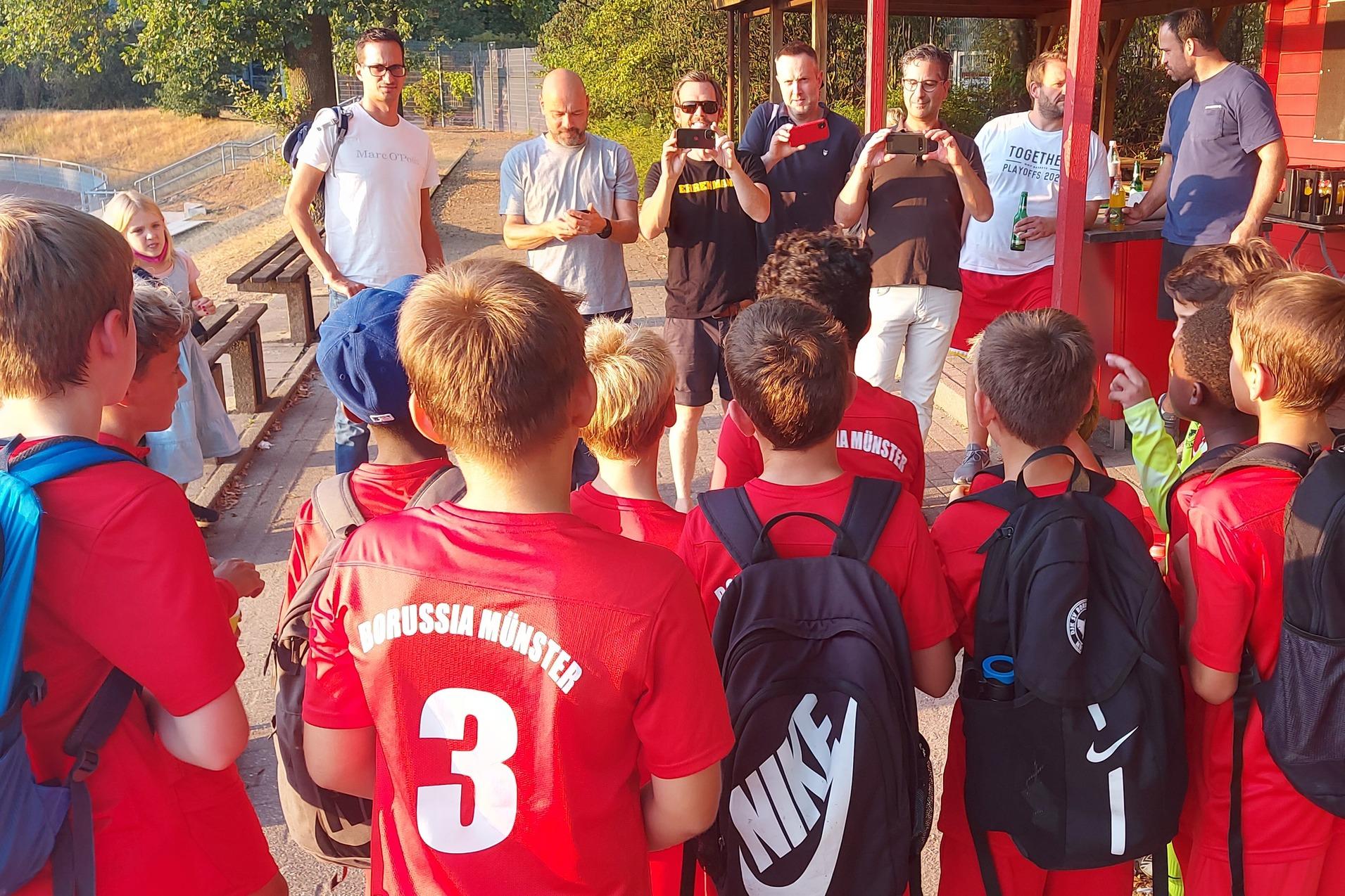 Gruppe von Kindern im roten Fußballtrikot hört einem Trainer an einem Vereinsheim zu.