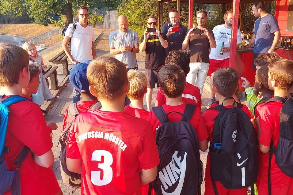 Gruppe von Kindern im roten Fußballtrikot hört einem Trainer an einem Vereinsheim zu.