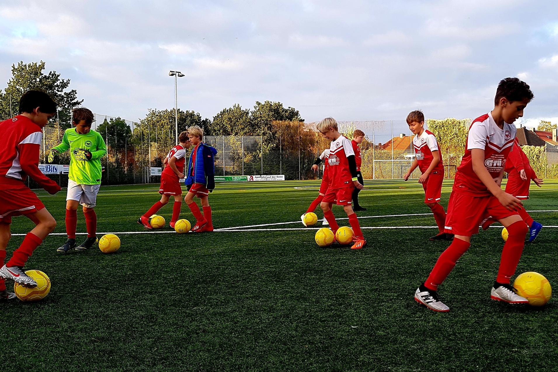 Jugendliche Fußballspieler in roten Trikots üben mit gelben Bällen auf einem Kunstrasenplatz unter bewölktem Himmel.