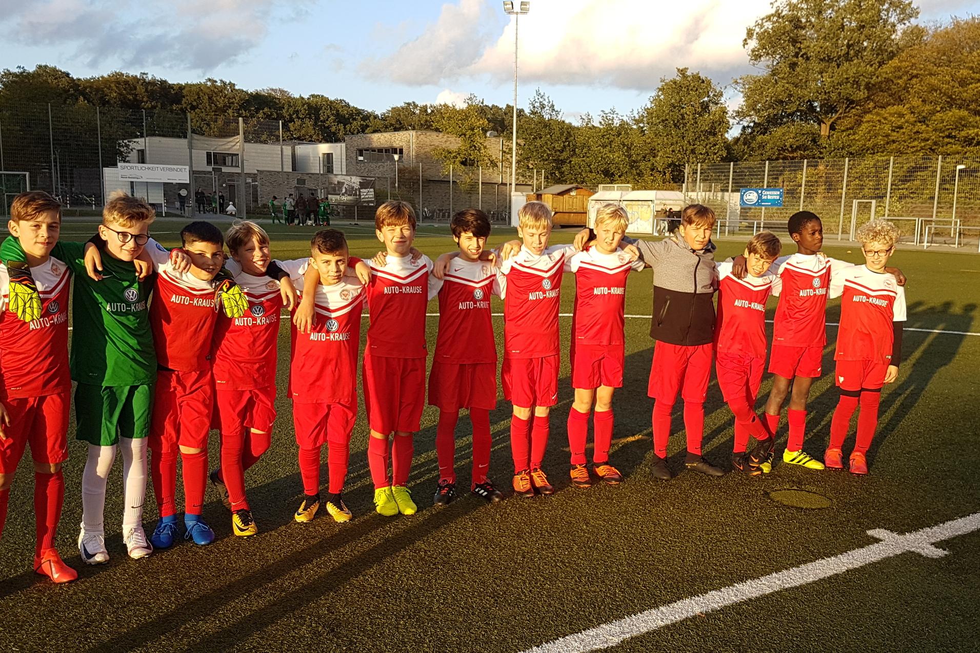 Gruppe von zehn jungen Fußballspielern in roten Trikots, auf einem Fußballfeld stehend, mit Sportplatz im Hintergrund.