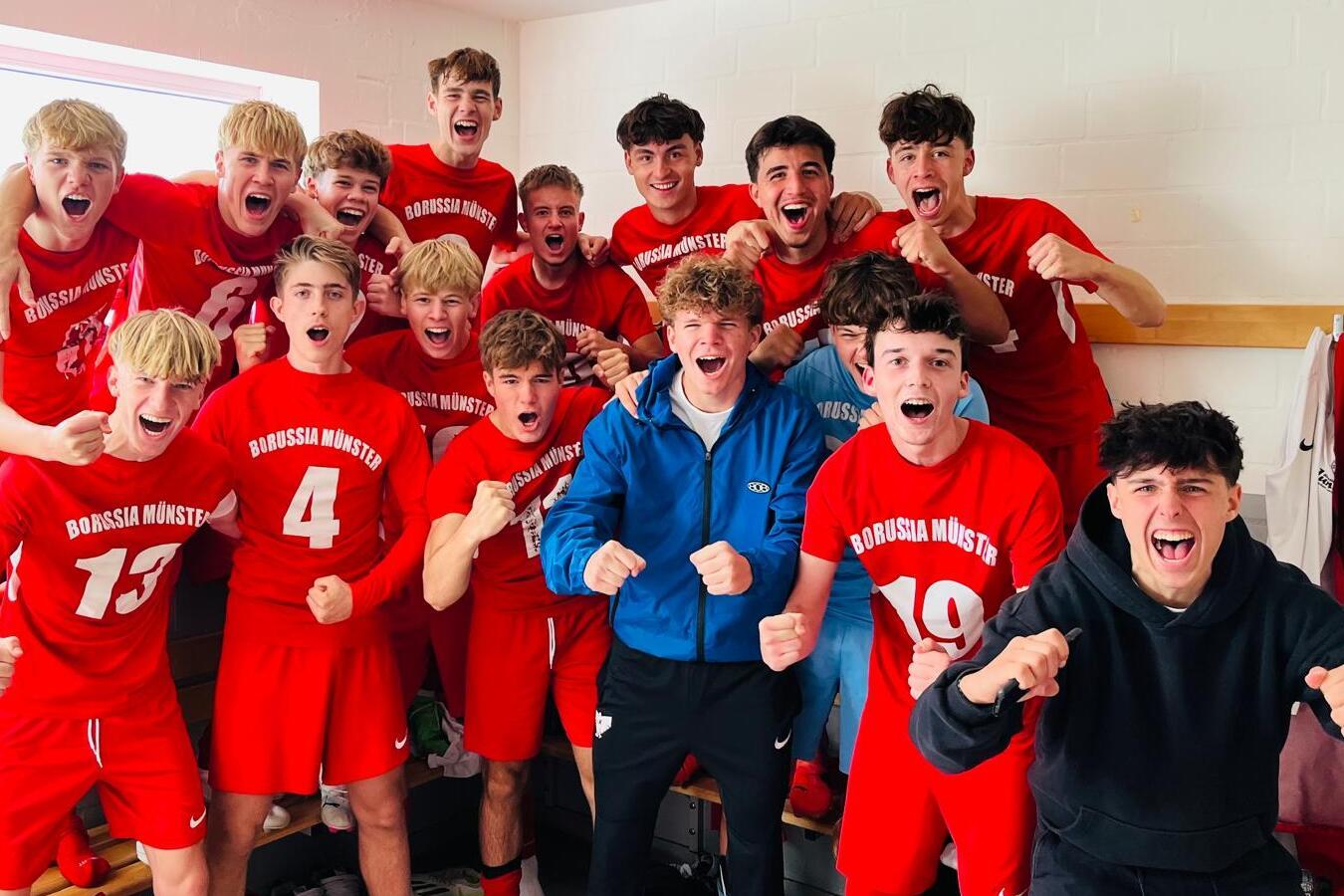 Gruppe von jubelnden jungen Fußballspielern in roten Trikots, die in einer Umkleidekabine stehen.