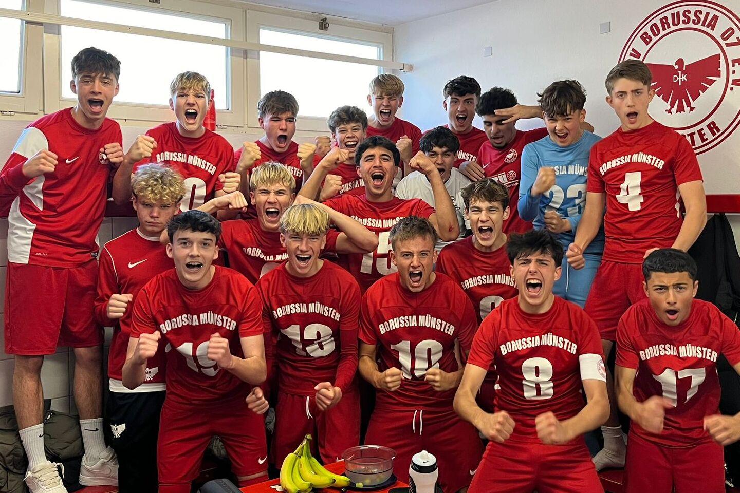 Gruppe junger Fußballspieler in roten Trikots jubeln fröhlich in einem Umkleideraum.