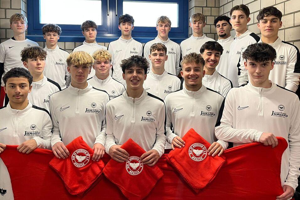 Gruppe von 16 jungen Fußballspielern in weißen Trikots mit roten Flaggen, aufgereiht in einem Umkleideraum.