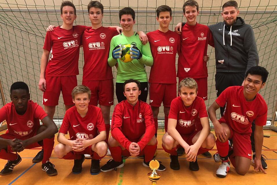 Jugendfußballmannschaft in roten Trikots posiert mit Pokal vor einem Tor in einer Sporthalle.