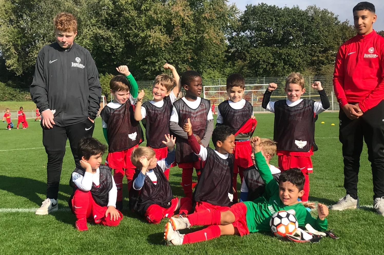 Gruppe von Kindern in Fußballtrikots posiert lächelnd auf einem grünen Sportplatz, zwei Trainer stehen hinter ihnen.