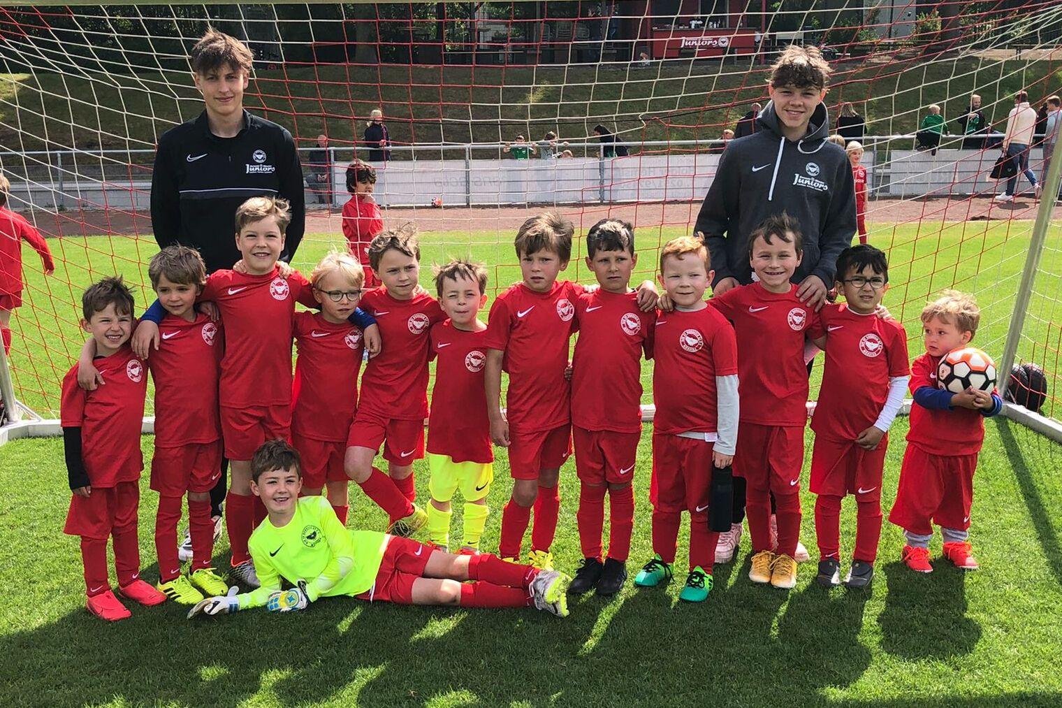 Gruppe von 14 Kindern in roten Fußballtrikots und zwei Trainern vor einem Tor auf einem Fußballfeld.