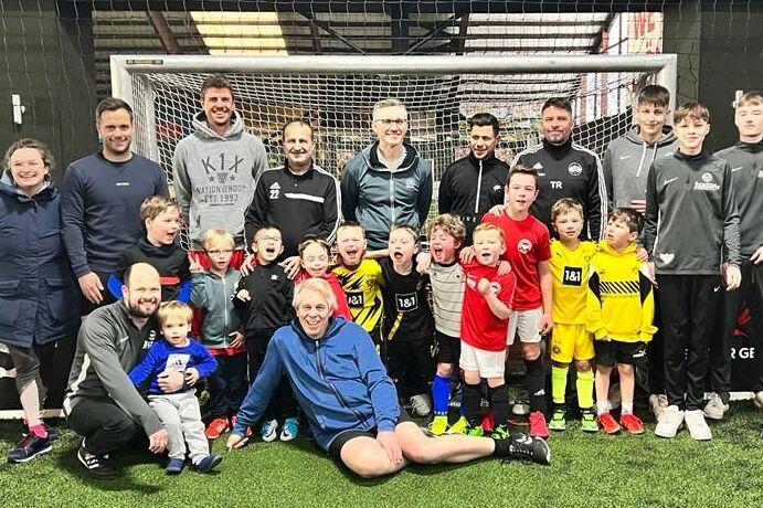 Gruppenfoto von Trainern und Kindern in Sportkleidung auf einem Fußballplatz, umgeben von einem Tor und Netz.