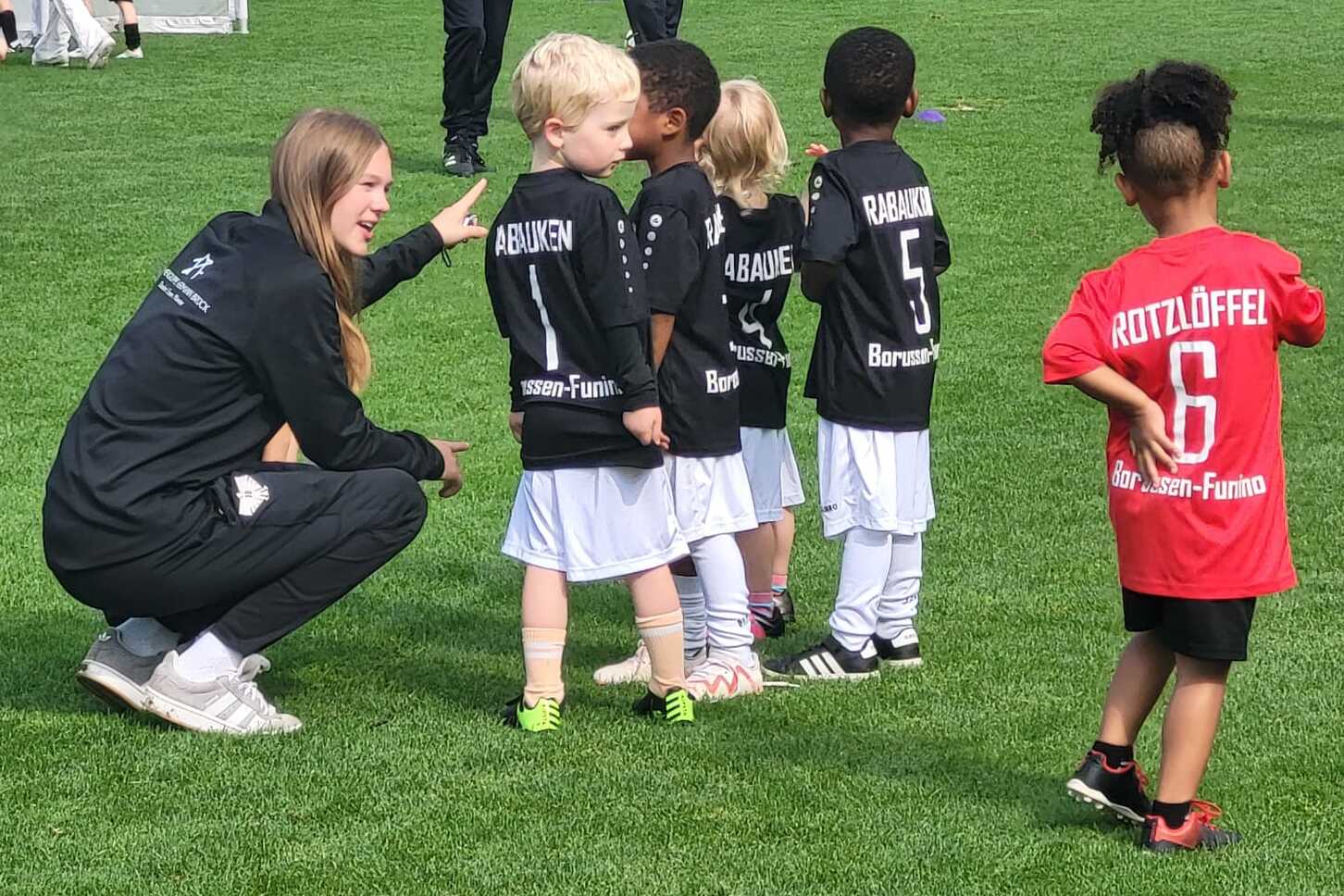 Trainerin spricht mit einer Gruppe von Kindern in Fußballtrikots auf einem Sportplatz.