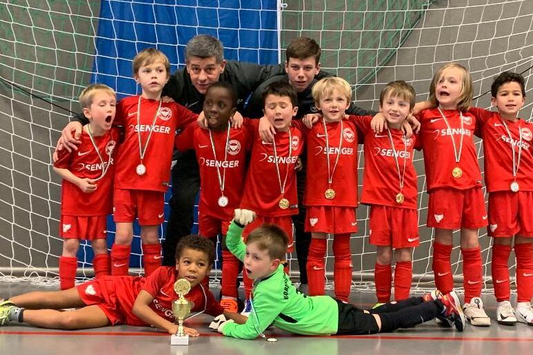 Eine Gruppenaufnahme von zwölf jubelnden Kindern in roten Fußballtrikots und zwei Trainern vor einem Tor, mit Medaillen.