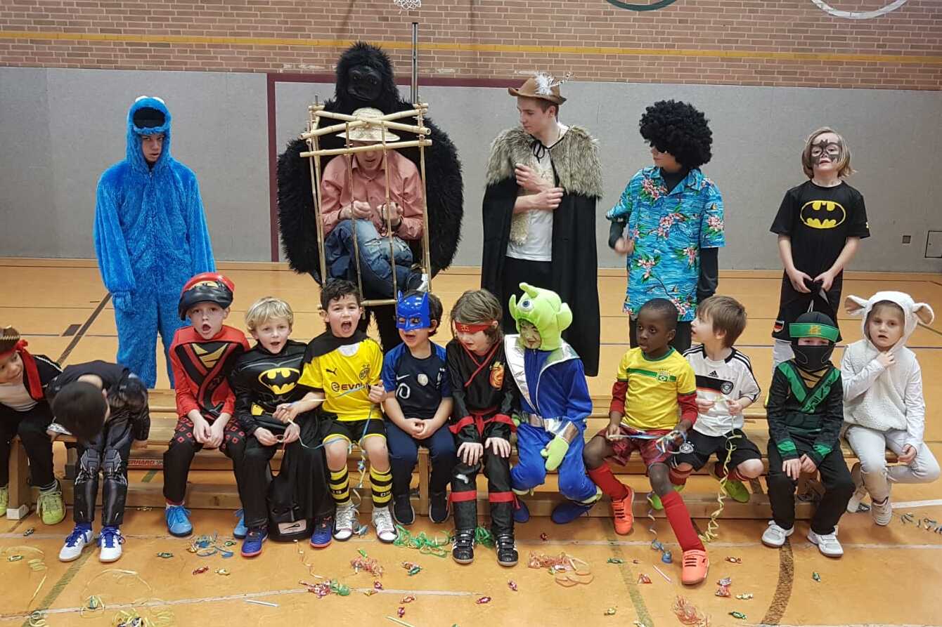 Gruppe von Kindern in Kostümen, die in einer Halle auf einer Bank sitzen, mit Konfetti auf dem Boden.