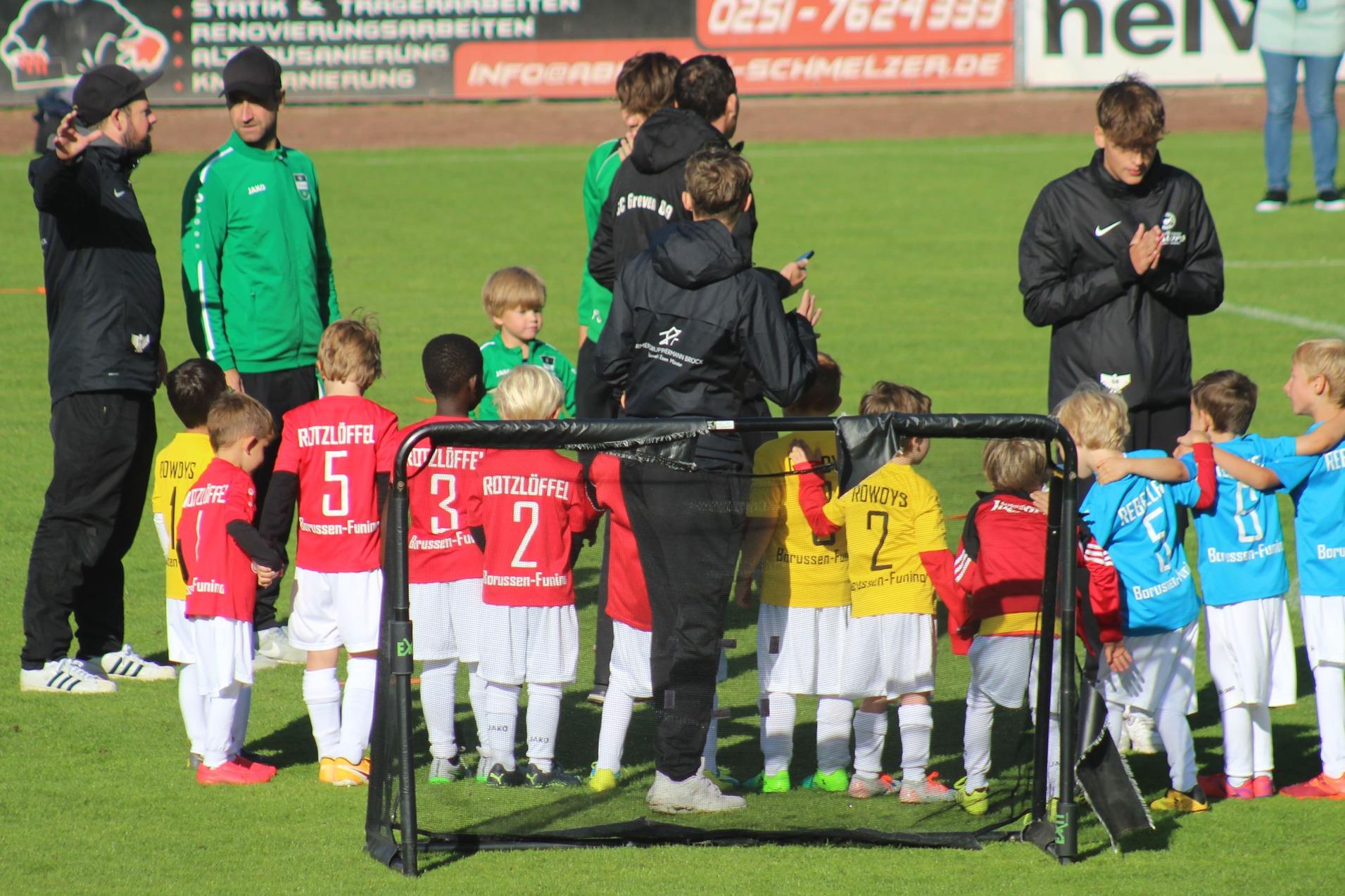 Zwei Gruppen von Kindern in Fußballtrikots stehen nebeneinander auf einem Rasenplatz, umgeben von Trainern.