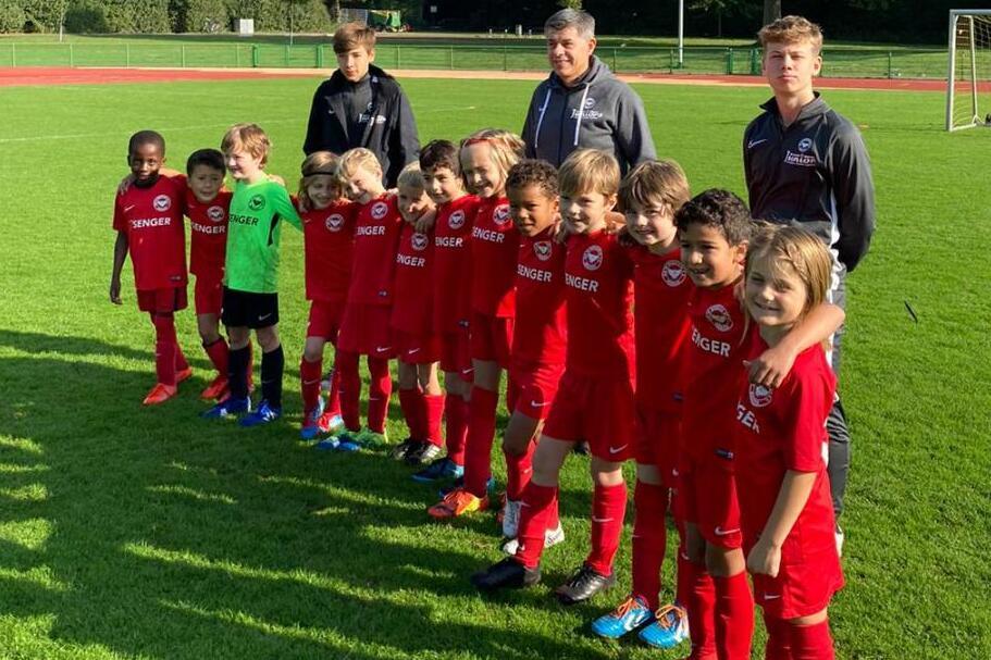 Gruppe von Kindern in roten Fußballtrikots mit Trainern auf einem Sportplatz, umgeben von Bäumen und einem Tor.