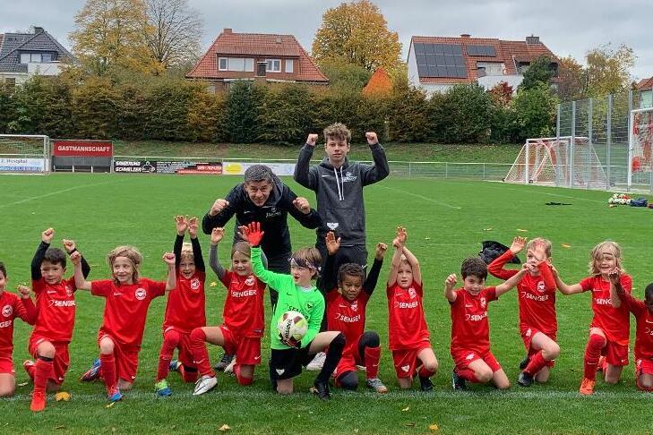 Gruppe von Kindern in roten Fußballtrikots posiert jubelnd auf einem Rasenfeld mit Toren im Hintergrund.