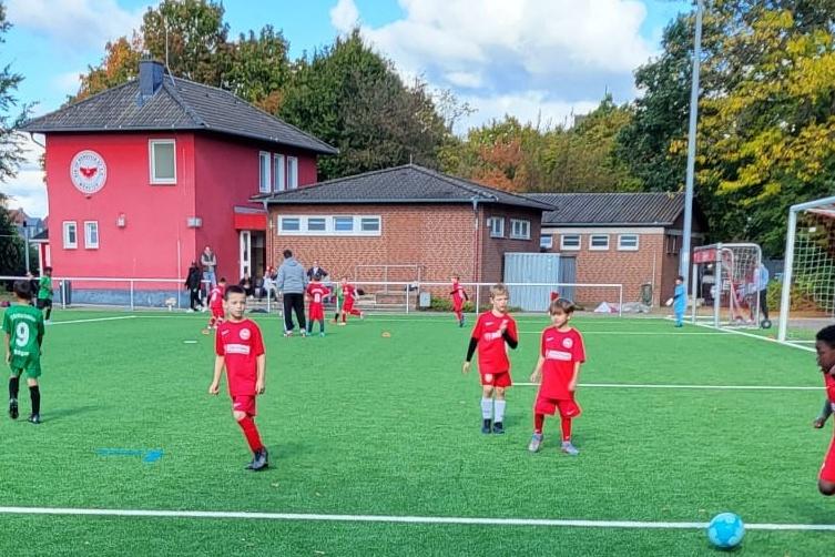 Sechs Kinder in roten und grünen Trikots spielen Fußball auf einem Kunstrasenplatz vor einem Vereinsgebäude.