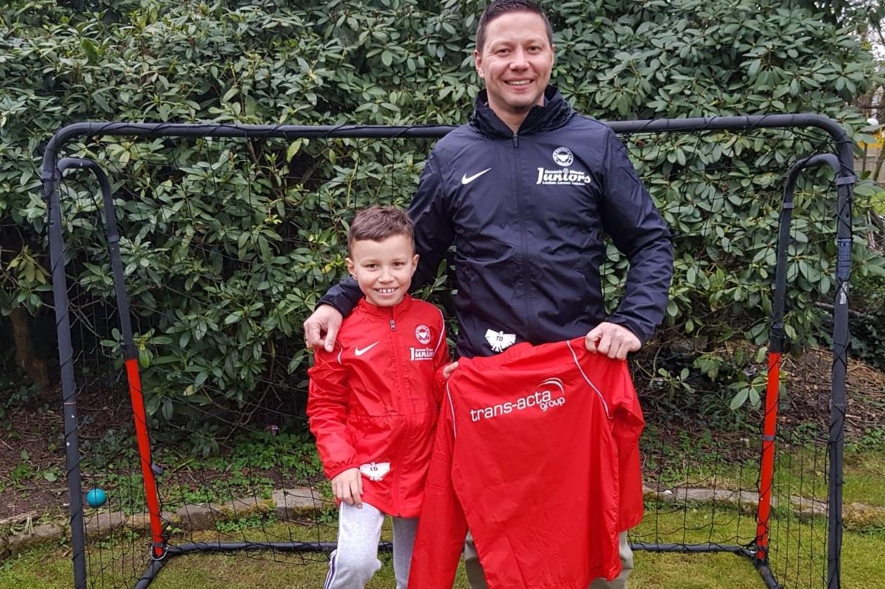Vater und Sohn stehen vor einem Fußballtor, beide in roten Trainingsjacken, der Sohn hält ein Fußballtrikot in der Hand.