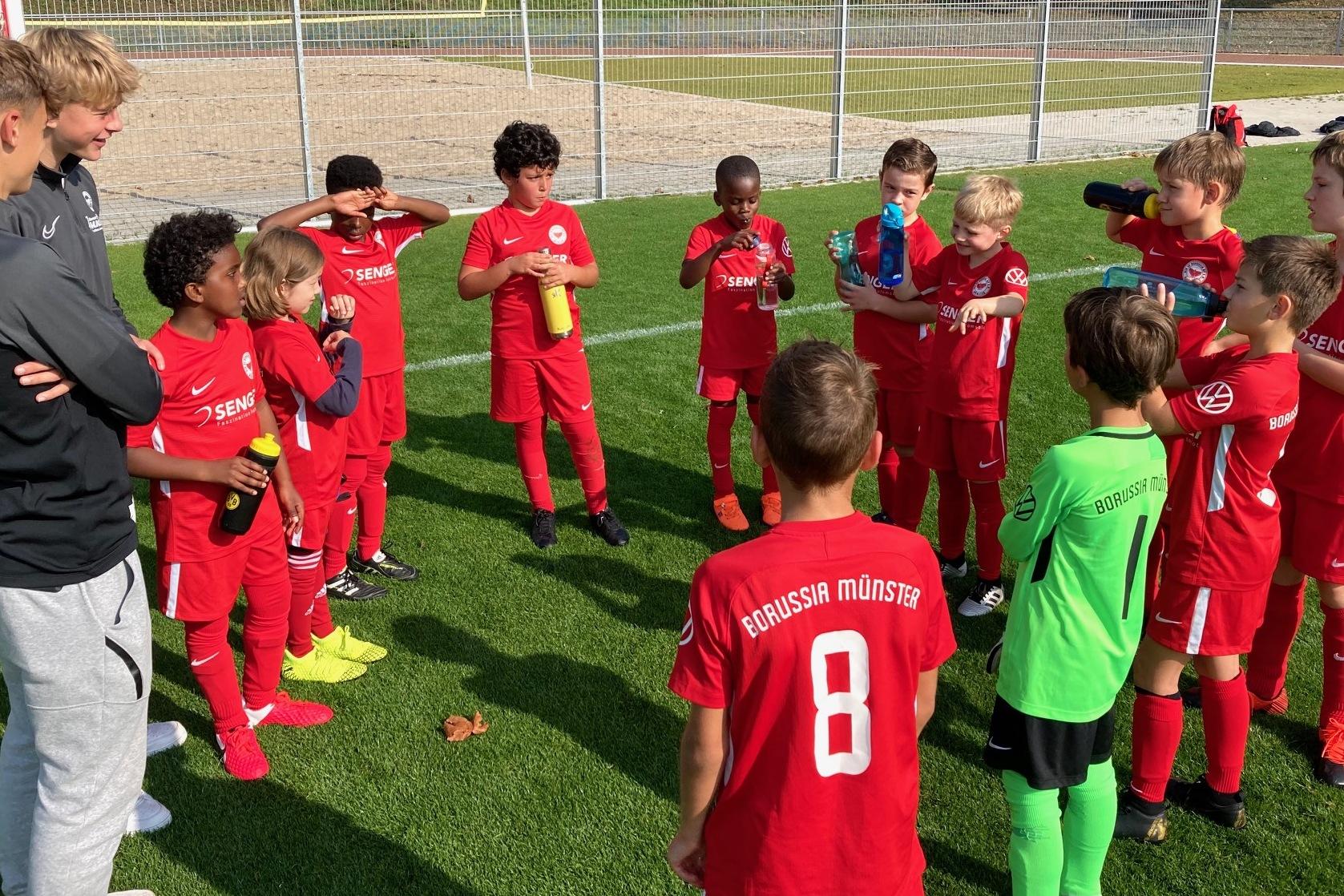 Gruppe von jungen Fußballspielern in roten Trikots versammelt sich um einen Trainer auf einem Sportplatz.