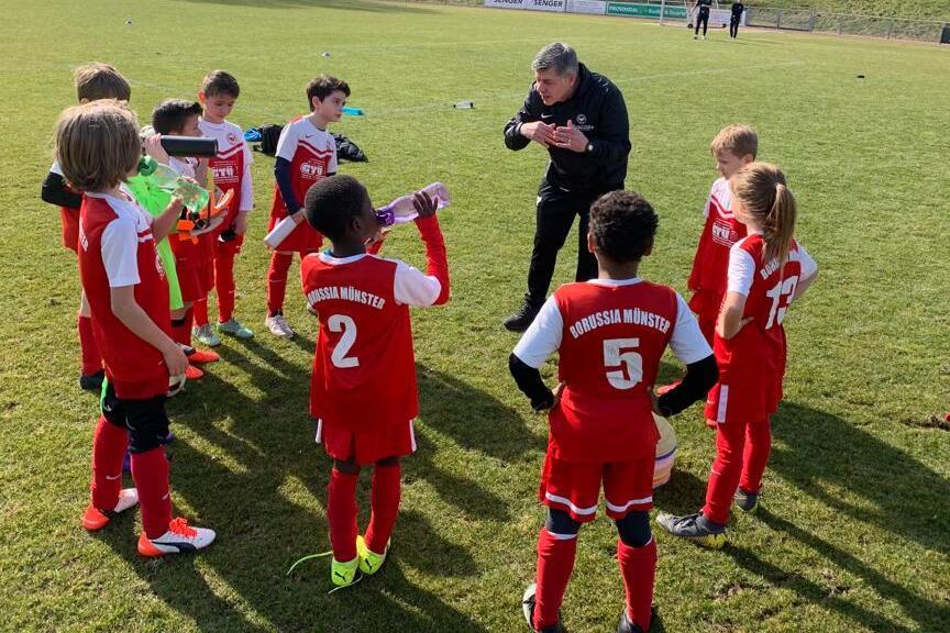 Trainer spricht mit einer Gruppe junger Fußballspieler in roten Trikots auf einem Rasenfeld.