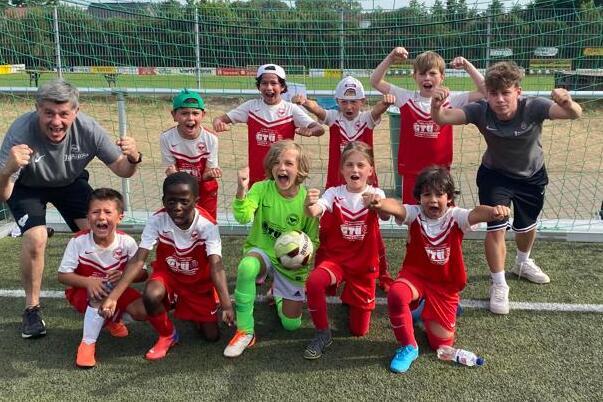 Gruppe von Kindern in roten Fußballtrikots, lachend und jubelnd vor einem Tor auf einem Kunstrasenfeld.