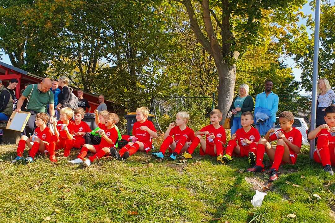 Gruppe von Kindern in roten Fußballtrikots sitzt auf einer Wiese, Zuschauer im Hintergrund. Straße im Bild beschildert.