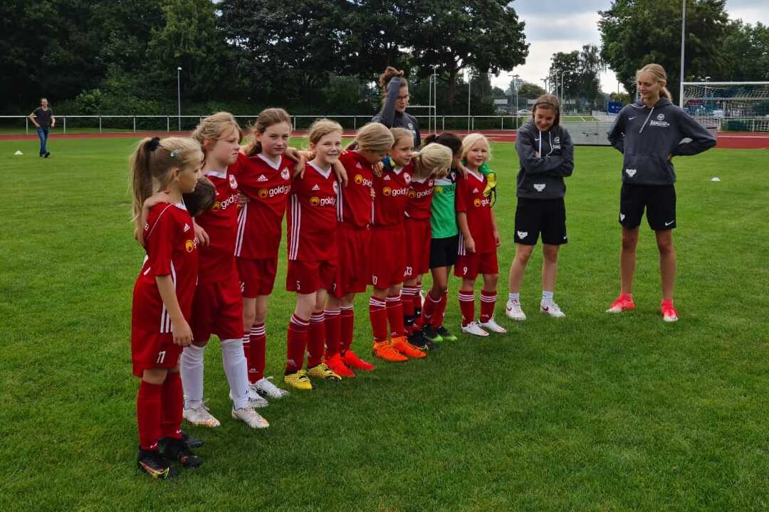 Gruppe junger Fußballspielerinnen in roten Trikots steht auf einem Rasenplatz, beobachtet von Trainern.