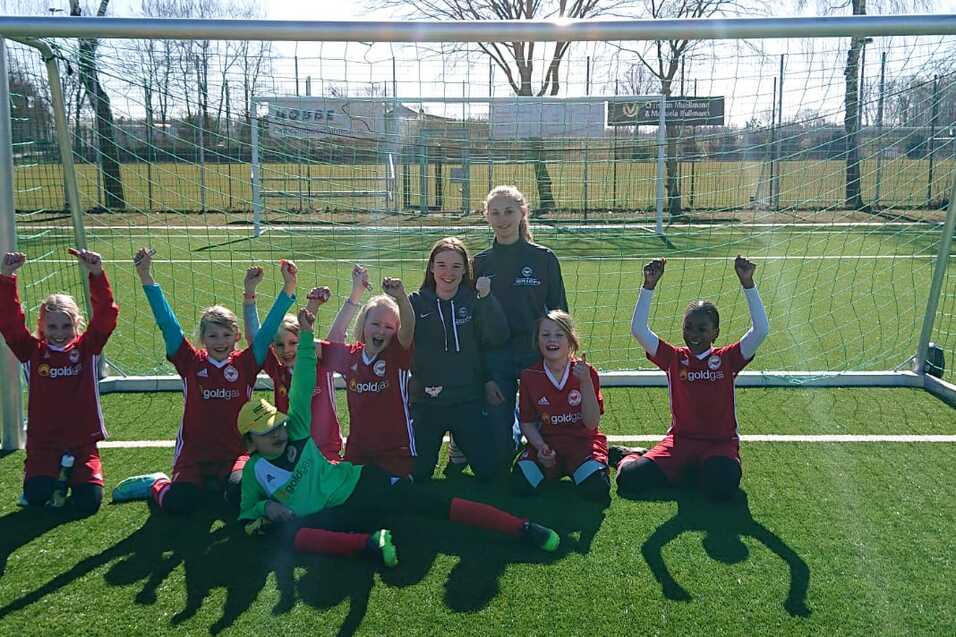Sechs junge Fußballspielerinnen posieren glücklich vor einem Tor auf einem Kunstrasenplatz unter klarem Himmel.